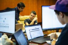 UVA students seated around a conference room table covered with computers, computer monitors, and paper and pencils, work intently during the qualifying round  of the Mid-Atlantic Collegiate Competition.