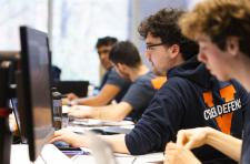 UVA students seated around a conference room table covered with computers, computer monitors, and paper and pencils, work intently during the qualifying round  of the Mid-Atlantic Collegiate Competition.