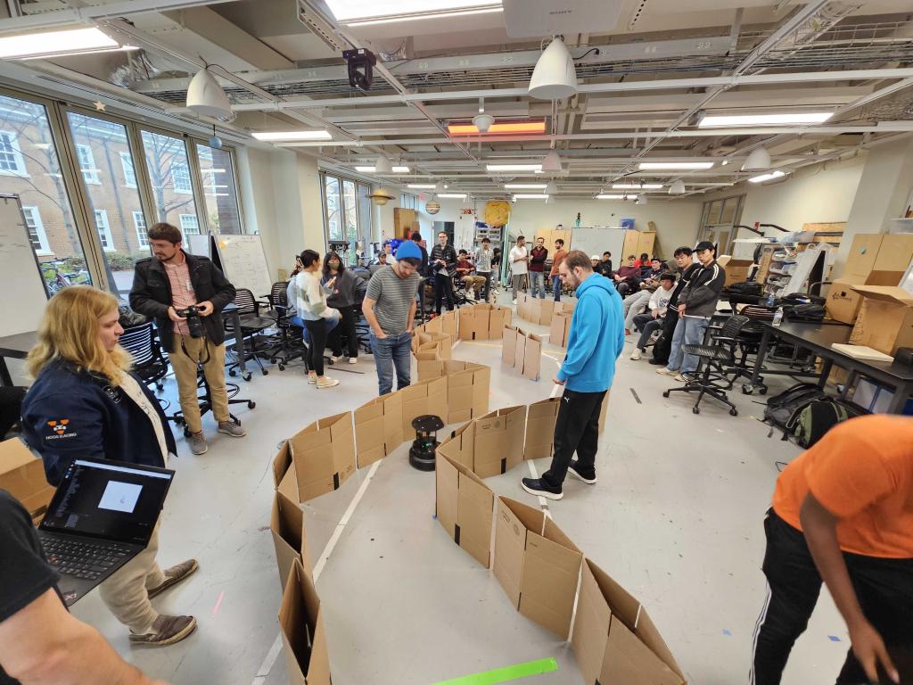A robot navigates a maze set-up with cardboard on the floor of a large room with students standing around watching. 