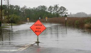 A road sign that reads "Caution High Water" with a flooded street in the background