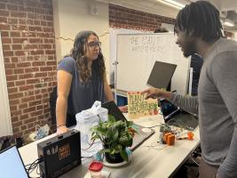 Two students talking by a table with a plant and "plant whisperers" group name sign