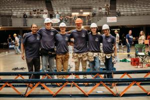Instructor José Gomez and students wearing hard hats pose behind the steel bridge at the competition.