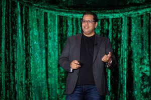 Madhur Behl delivers his keynote speech in front of a shiny green curtain.