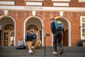 A student sits with a laptop on Thornton Hall steps as another student passes.